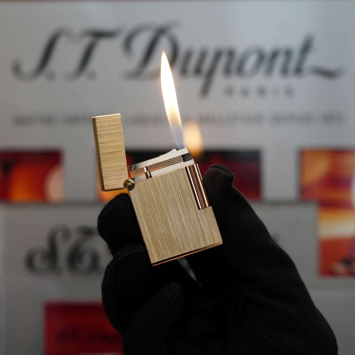 A gloved hand holds an open gold lighter with a lit flame in front of a blurred background, showcasing the "S.T. Dupont" branding. This exquisite piece from the Vintage 1980 St Dupont Gold brushed finish Medium Ligne 2 lighter collection exemplifies timeless elegance and craftsmanship.