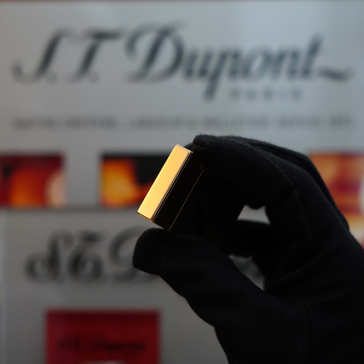 A gloved hand holds a small, gold-colored rectangular object from the Vintage 1980 St Dupont Gold brushed finish Medium Ligne 2 lighter series in front of a blurred background featuring the text "S.T. Dupont," capturing the essence of a true collector's item.