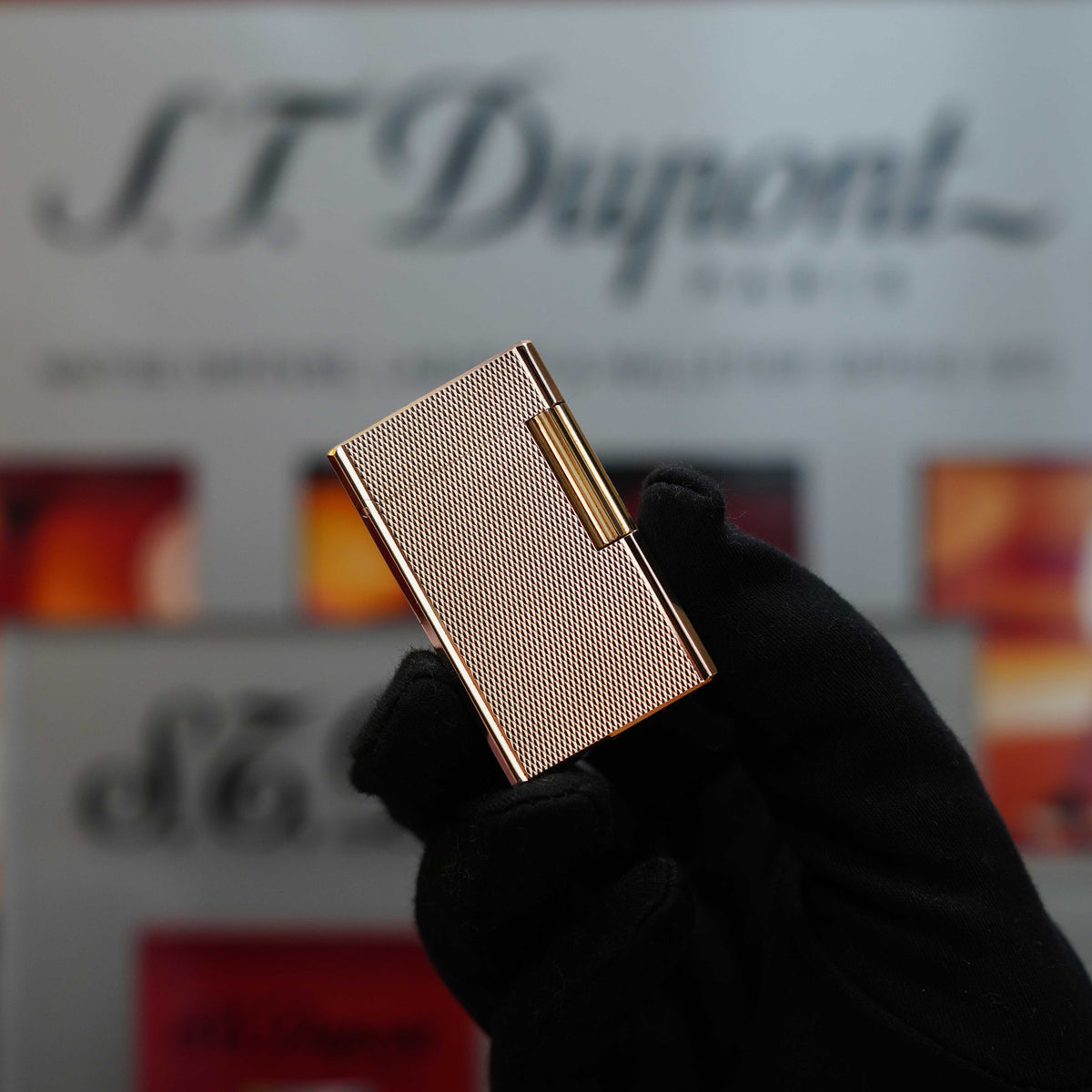 A gloved hand holds a Vintage 1970 St Dupont Micro Diamond rare Rose Gold Large Ligne 1 lighter in front of a blurred S.T. Dupont display background, showcasing this collector's item.