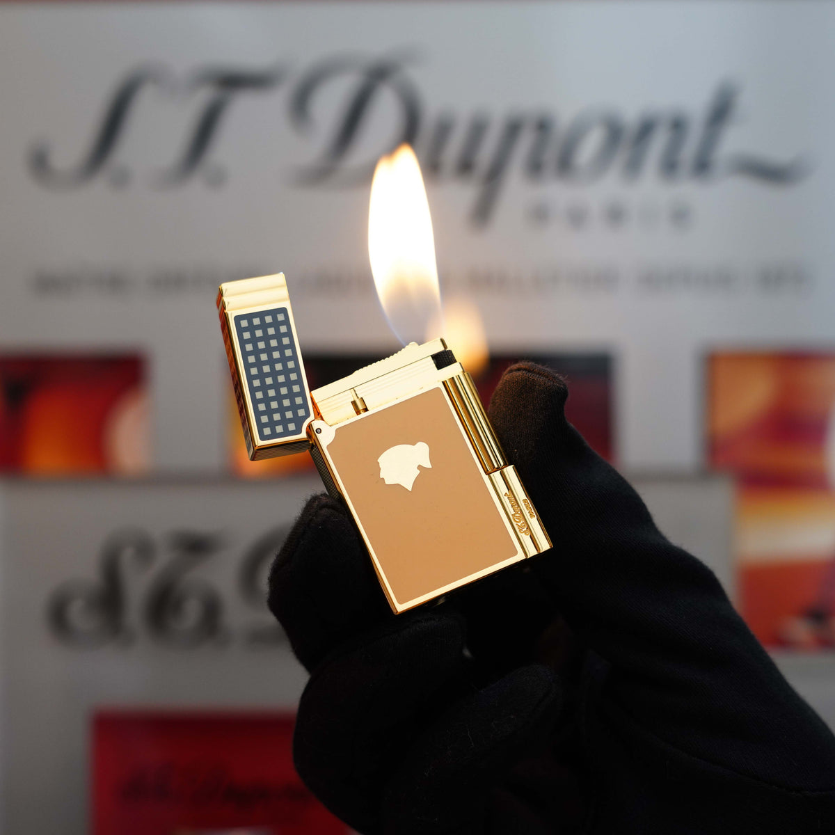 A hand in a black glove holding a lit, Vintage 1992 rare St Dupont Cohiba First Edition Dual Flame Gatsby 24k Gold finish natural Lacquer lighter with a rectangular grid design on the lid in front of a blurred background featuring "S.T. Dupont" text.
