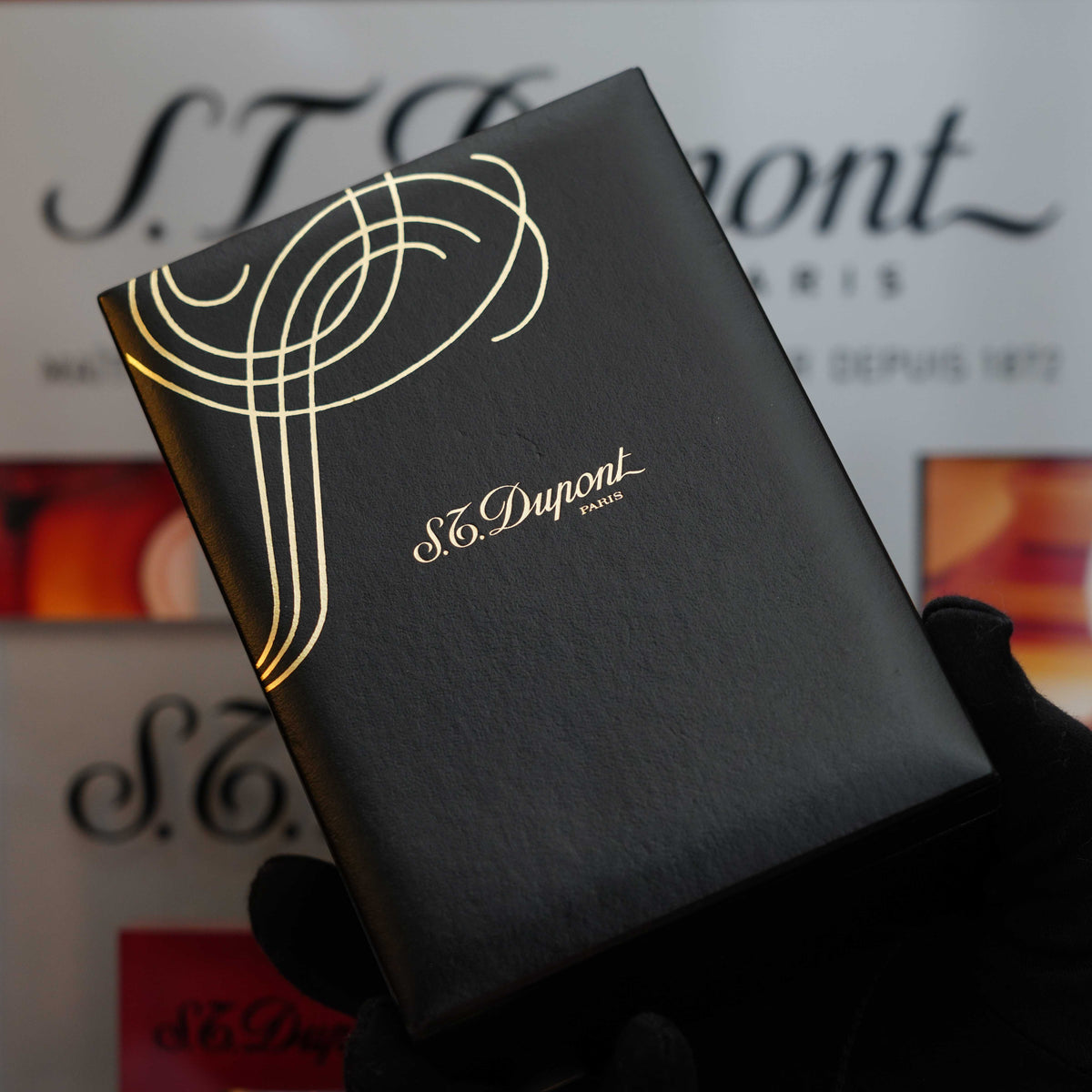 A person wearing black gloves holds an S.T. Dupont Paris box with gold lettering and design, showcasing a Vintage 1993 rare Art Nouveau early Limited Editions 24k Gold finish natural Green lacquer ligne 2 lighter, in front of a blurred background featuring S.T. Dupont branding.