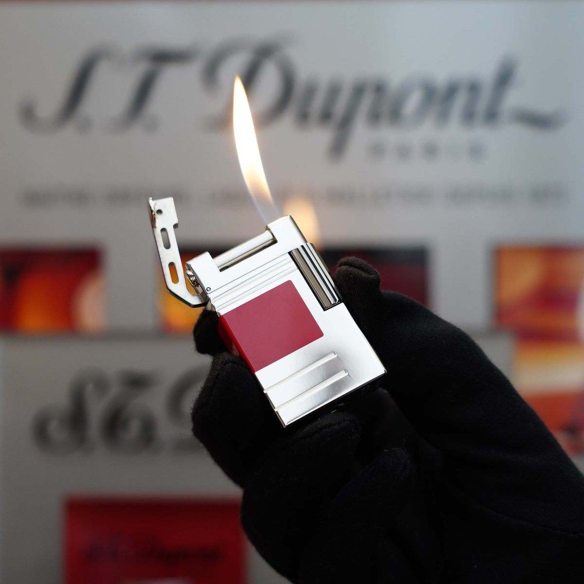 A gloved hand holds a lit Vintage 1999 S.T. Dupont Urban Limited Edition Platinum finish natural Red Lacquer lighter, an exquisite collector's item, in front of a blurry S.T. Dupont logo background.