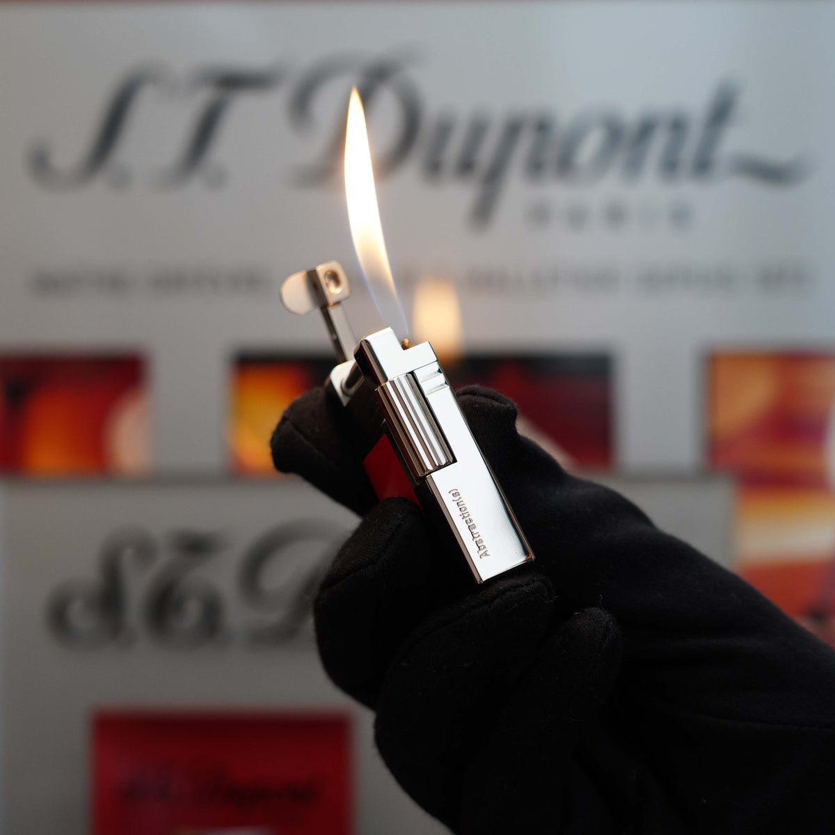 A person wearing a black glove holds a lit Vintage 1999 St Dupont Urban Limited Edition Platinum finish natural Red Lacquer lighter, its flame dancing against a blurred background featuring S.T. Dupont branding.