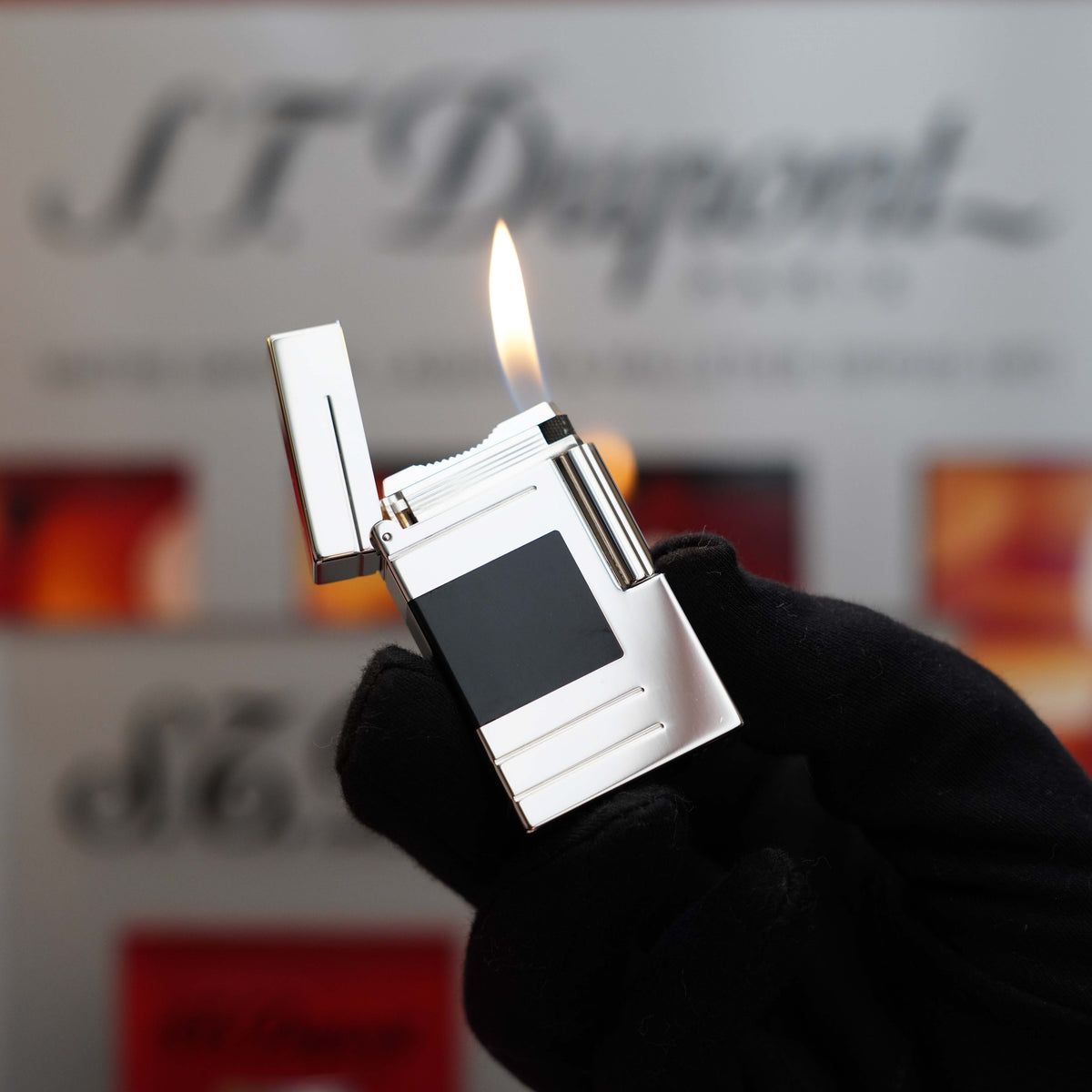 A person wearing a black glove holds a lit silver cigarette lighter with a black panel, showcasing the "S.T. Dupont" logo visible in the blurred background—a Vintage 1999 St Dupont Ligne 1 Limited Edition Platinum finish natural Black Lacquer lighter that exudes timeless elegance.