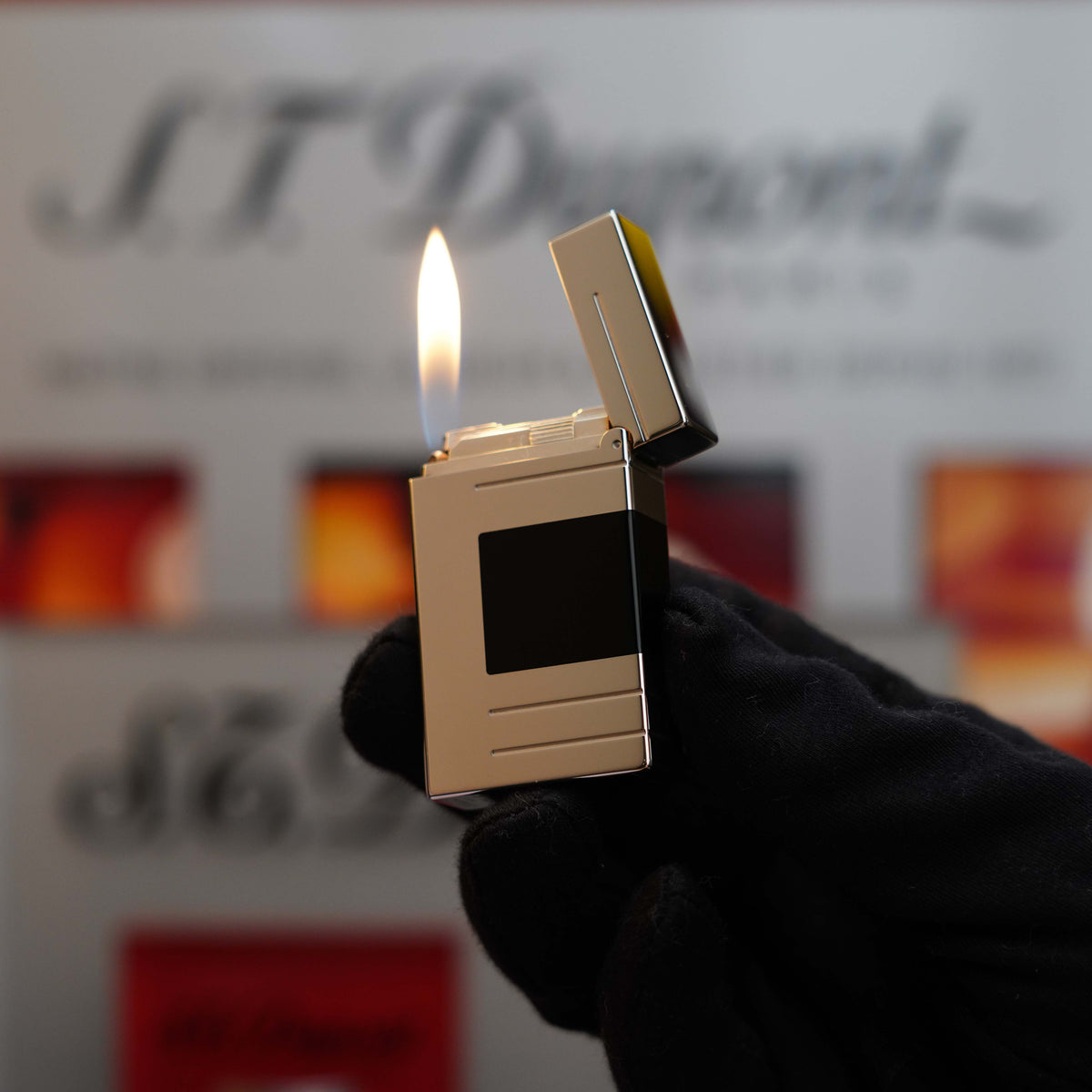 A hand in a black glove holding a lit, Vintage 1999 S.T. Dupont Ligne 1 Limited Edition Platinum finish natural Black Lacquer lighter with a black square on it, against a blurred background featuring text and images.