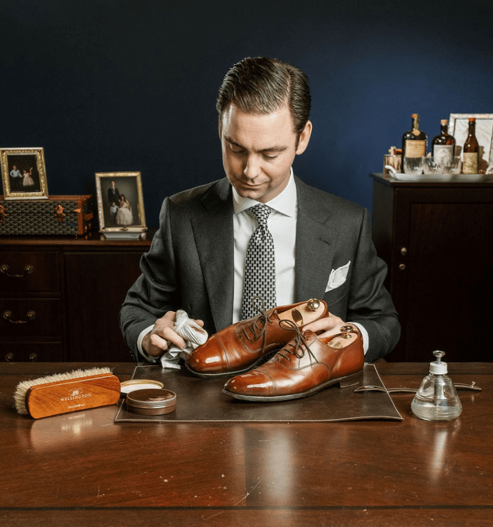 Image of Kirby Allison, sitting at a wooden desk looking down while polishing a pair of brown loafers with a cloth.