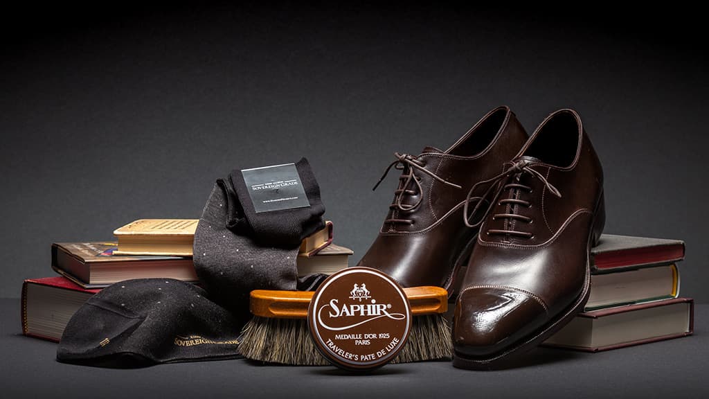 Photo of a pair of brown dress shoes, a Saphir shoe polish can, black dress socks, and shoe polishing brush arranged over stacks of books. 