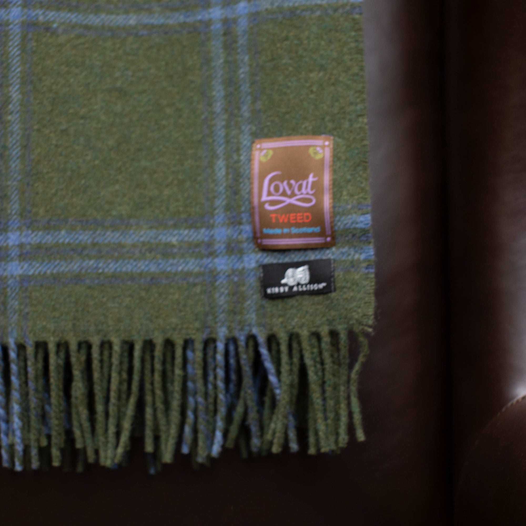 A green and blue plaid Kirby Allison Lovat Mill Suit to Shoot Tweed wool throw blanket on a leather chair.