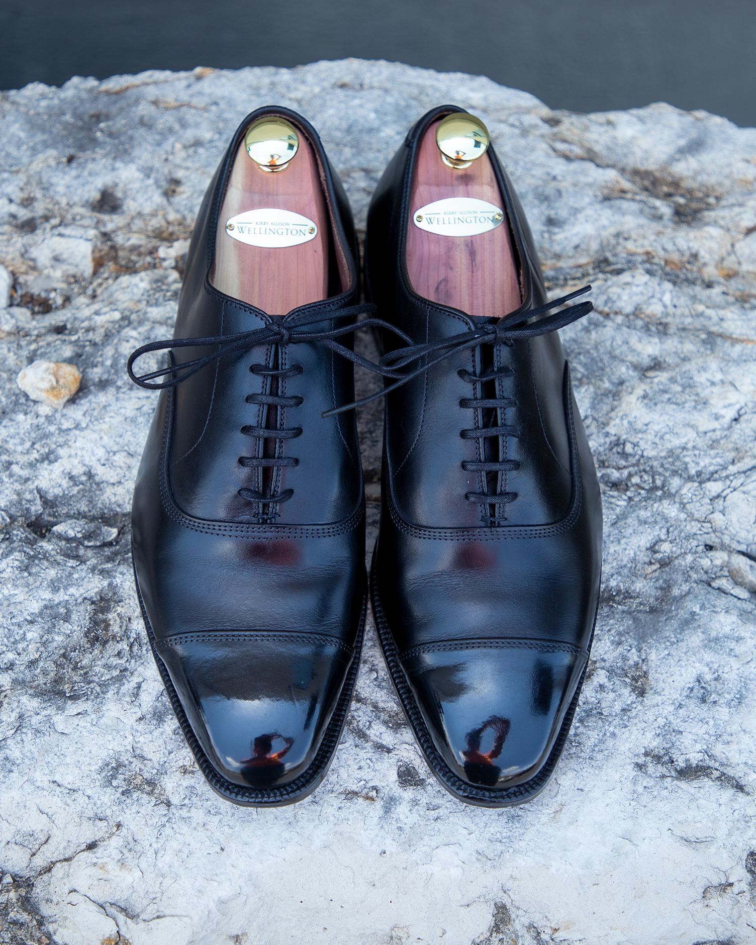 A pair of black oxford shoes with the KirbyAllison.com High Shine Service sitting on a rock.