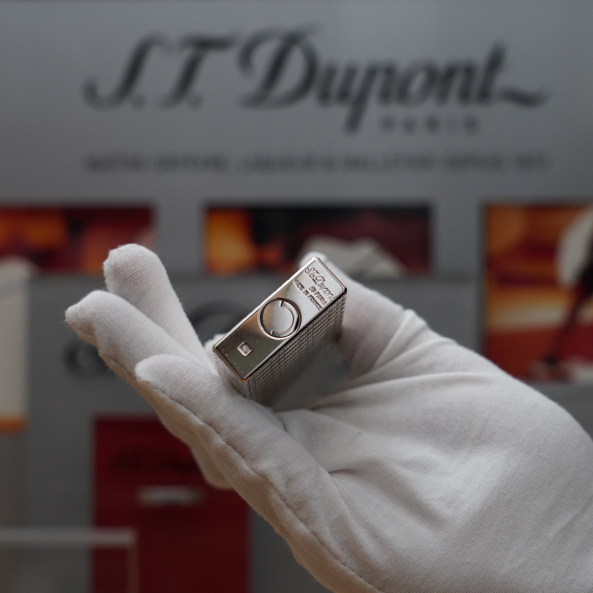 A Vintage 1970 S.T. Dupont Silver Plated Ligne 1 Rare lines pattern Table Lighter being held in front of S.T. Dupont by a hand.