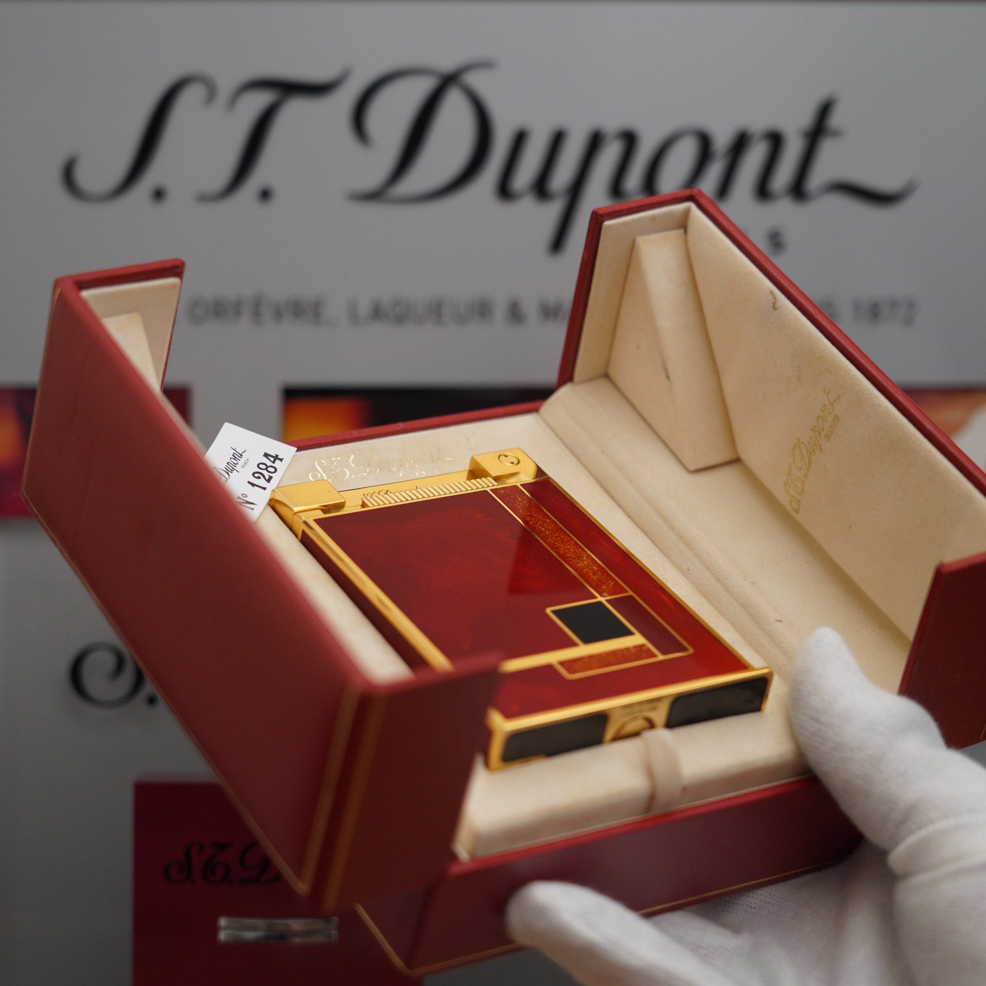 A person is holding a vintage 1990 S.T. Dupont 18k Jeroboam Gold Dust Red Chinese Lacquer Gold Plated Table Lighter inside a red box.