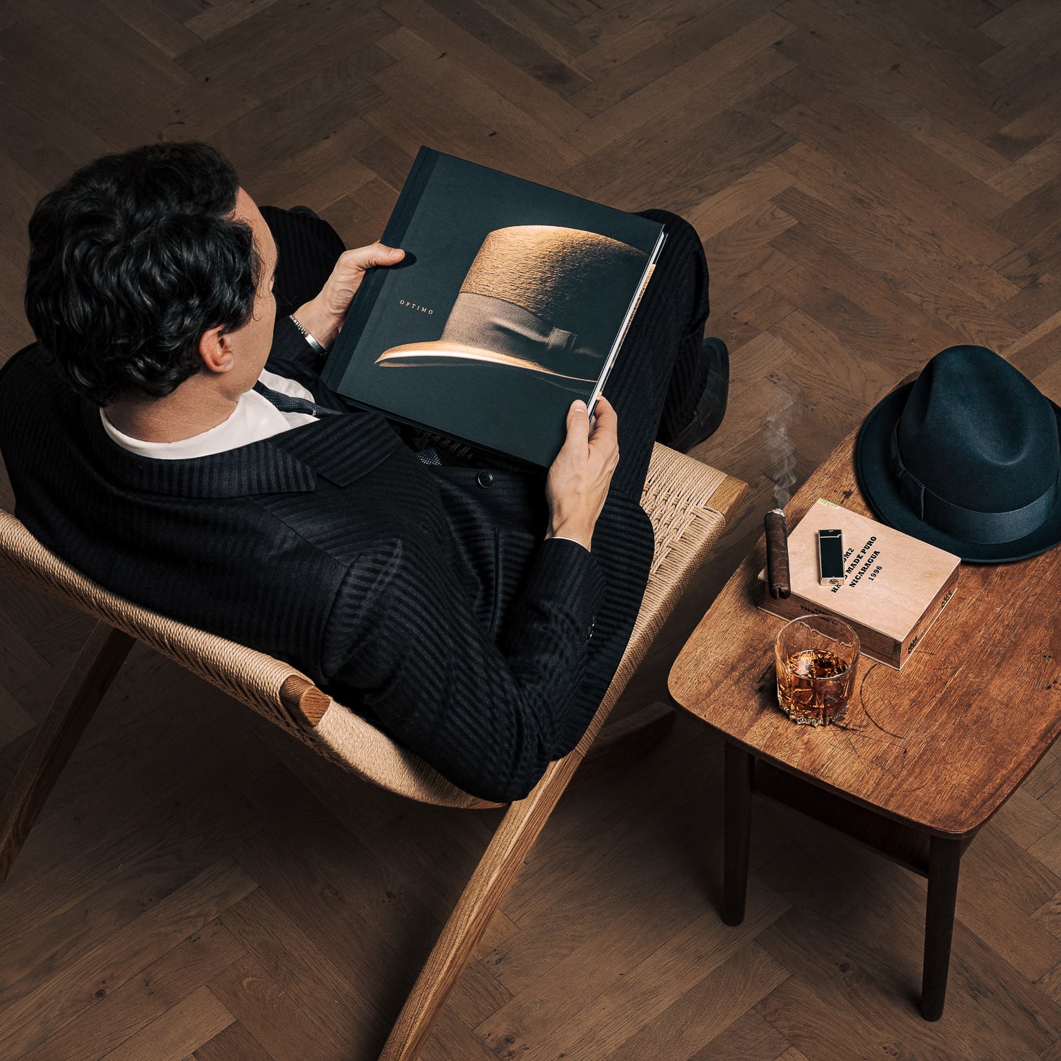 A man wearing the finest Optimo Hats: The Art of the Hatmaker sitting in a chair reading a book.