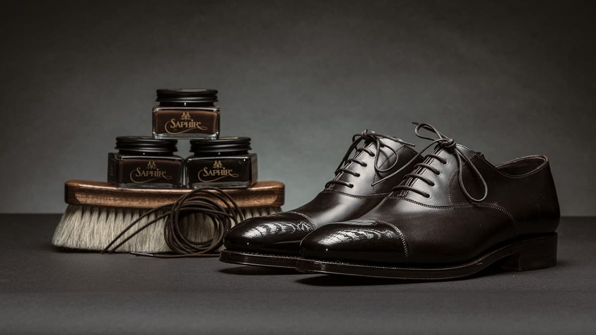 Image of a pair of black wing tip dress shoes arranged next to a three Saphir shoe polish jars stocked on a wood shoe polish brush with a coiled pair of laces leaning in front of the brush. 