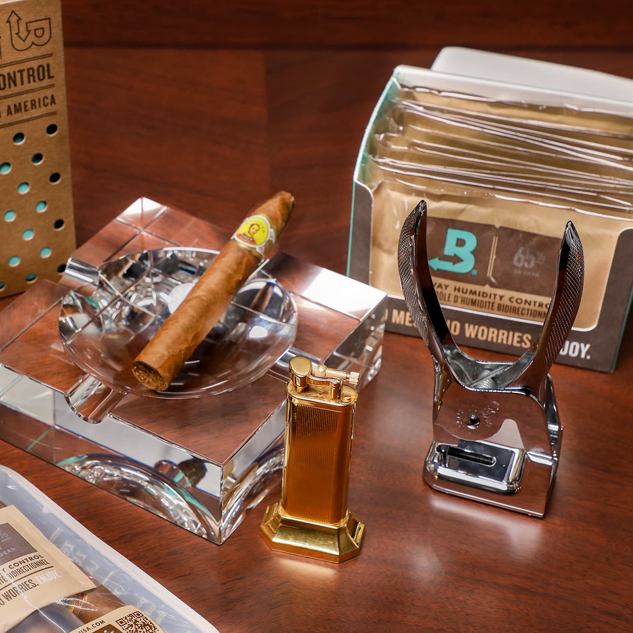 Close up image of a gold cigar lighter, a silver cigar cutter, and a glass ash tray with a cigar leaning on it, arranged on a brown wooden desk.