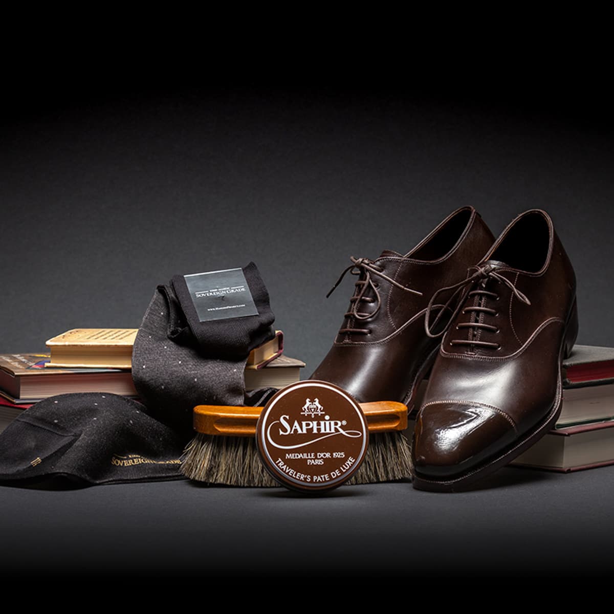 Image of a pair of brown dress shoes, a Saphir shoe polish can, black dress socks, and shoe polishing brush arranged over stacks of books. 