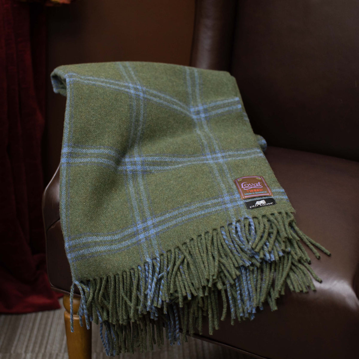 A new Lovat Mill Kirby Allison Suit to Shoot Tweed wool throw blanket in green and blue plaid, inspired by Scotland, on a chair.