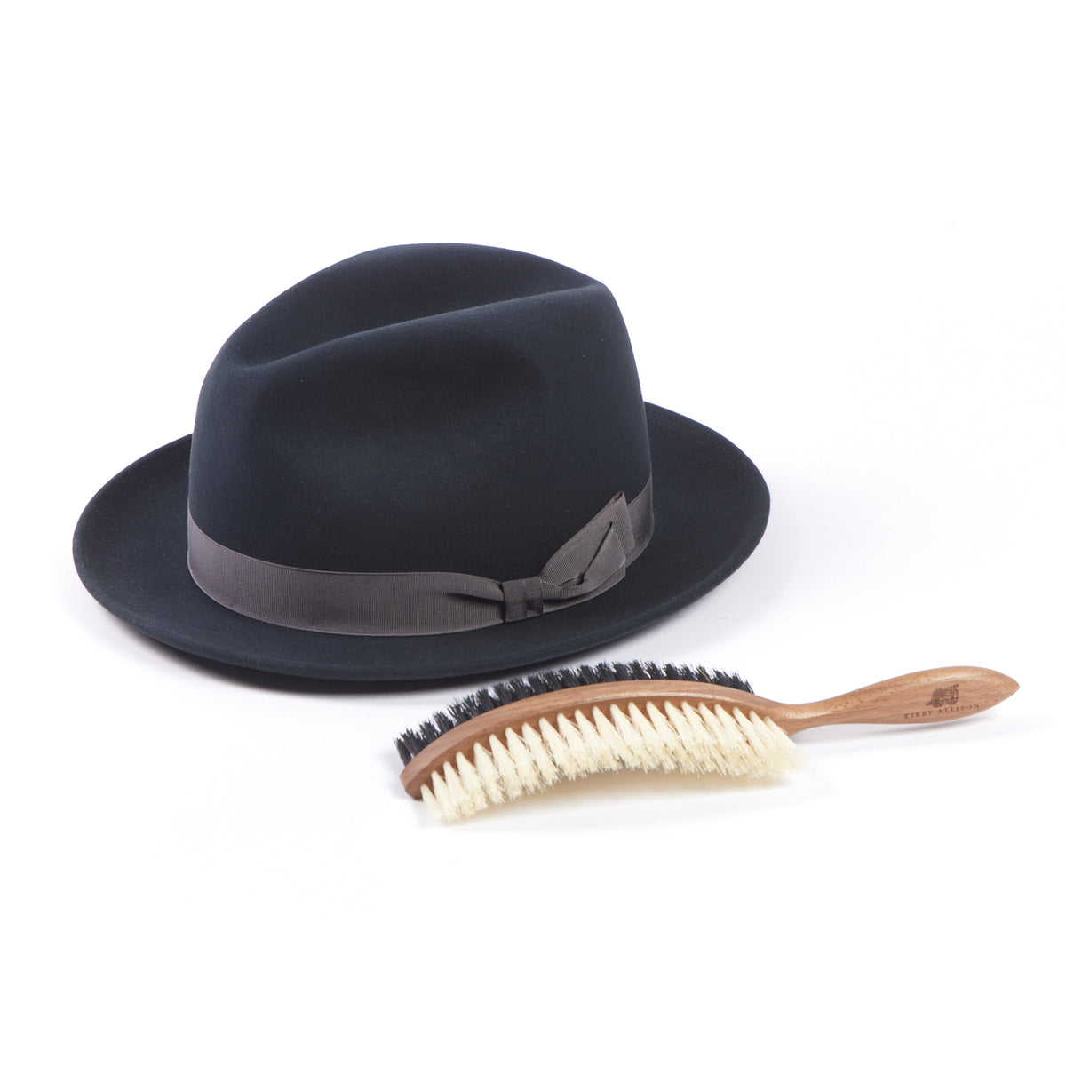 A black fedora hat and Kirby Allison Double-Sided Hat Brush on a white surface.
