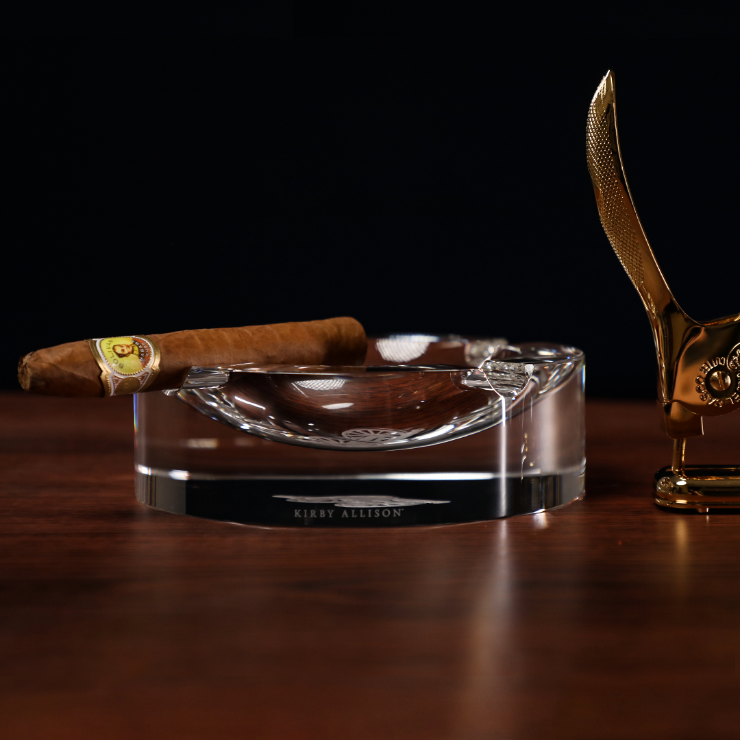 A smoking accessory featuring a Kirby Allison Glass Cigar Ash Tray - Round for finer cigars on a wooden table.