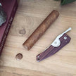 A friction folder knife with a stainless steel blade, the Kirby Allison Kingwood Cigar Knife from KirbyAllison.com, is displayed alongside a cigar on a wooden table.