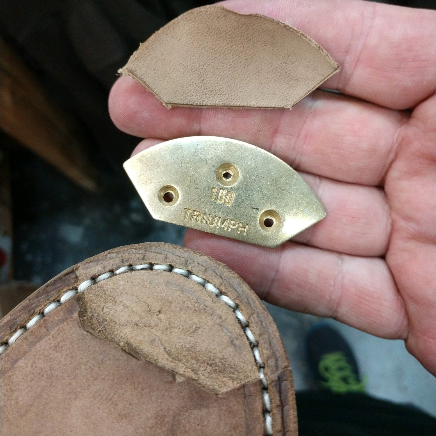 A person's hand holding a piece of leather with a KirbyAllison.com brass plate on it for the Toe Plate Only Service shoe restoration program.